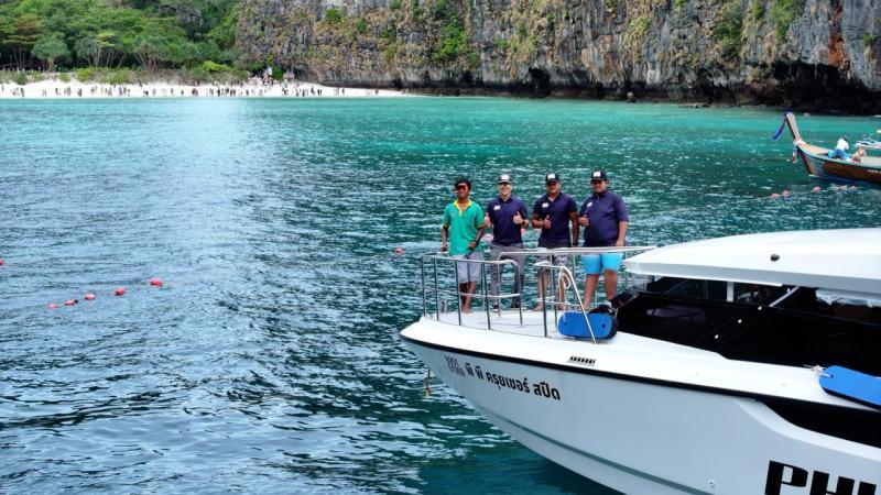 Four people on a boat near a tropical beach and cliffs.